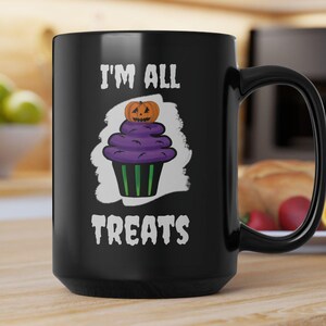May include: Black ceramic mug with a handle. The mug features a purple cupcake design with a pumpkin on top and the words "I'M ALL TREATS" in white. The mug is on a wooden surface.