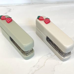 May include: Two staplers, one light green and one off-white, each adorned with two red strawberry embellishments with green leaves. The staplers are rectangular with a silver metal base, designed for office or school use.