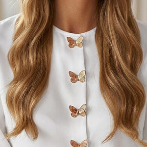 May include: A crisp white long-sleeved shirt featuring a vertical row of four metallic gold butterfly-shaped buttons down the center front placket. The shirt has a simple round neckline and a smooth, solid white fabric construction throughout.