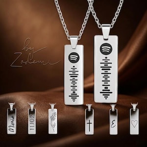 May include: Silver pendant necklaces with rectangular charms. The charms feature Spotify codes and hang from fine chains. Smaller pendants display text, a floral design, a cross, the Greek letter epsilon, and a heart.