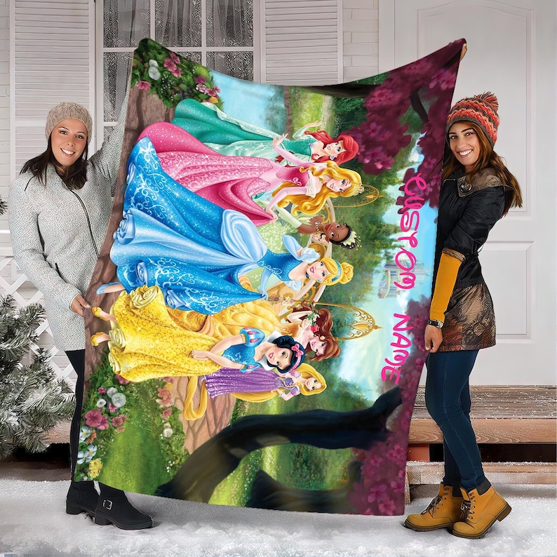 Puede incluir: Una colorida manta con ilustraciones de varias princesas de Disney con sus vestidos ic&oacute;nicos. La manta tiene un nombre personalizado impreso en el lateral. Las princesas est&aacute;n ambientadas en un bosque y un castillo.