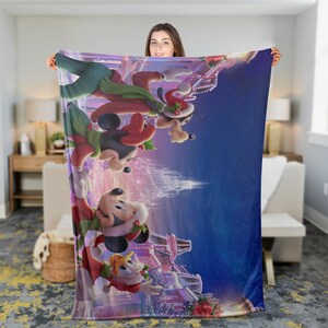 May include: A large, rectangular blanket featuring a festive holiday scene with animated characters. The blanket has a blue background with a colorful illustration of cartoon characters wearing Santa hats and holiday attire. The blanket is held up by a person.
