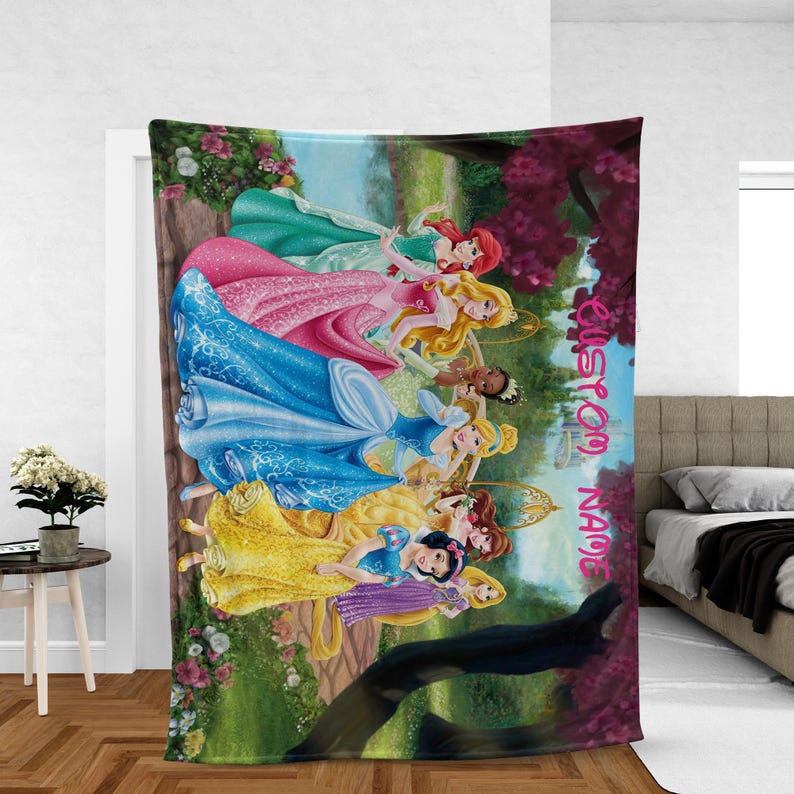Puede incluir: Una colorida manta con ilustraciones de varias princesas con sus vestidos ic&oacute;nicos. La manta tiene una combinaci&oacute;n de colores verde, rosa y azul con las palabras "CUSTOM NAME" en escritura rosa. Las princesas est&aacute;n ambientadas en un fondo de bosque.