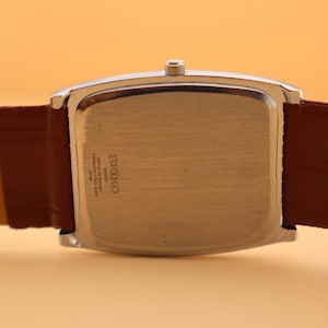 May include: A silver-toned rectangular watch case with a brown leather strap. The watch face is not visible. The back of the watch has the brand name "Seiko" and other text. The watch band is a medium brown color.