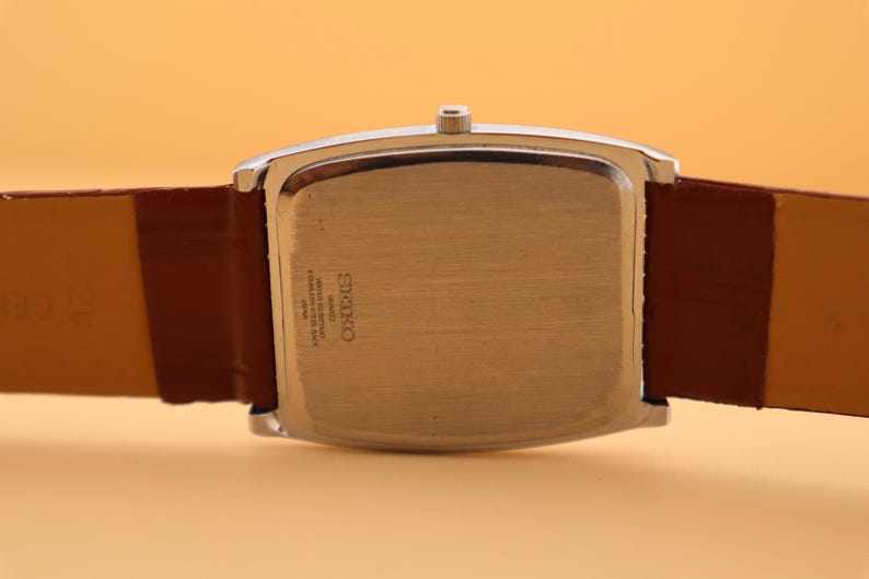 May include: A silver-toned rectangular watch case with a brown leather strap. The watch face is not visible. The back of the watch has the brand name "Seiko" and other text. The watch band is a medium brown color.