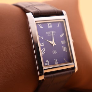 May include: A rectangular Seiko Quartz watch with a dark blue face, silver-tone case, and brown leather band. The watch face features Roman numeral hour markers and gold-tone hands. The watch is displayed on a brown leather pillow.