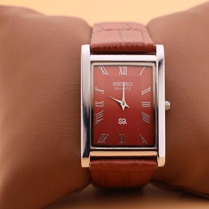 Könnte beinhalten: Eine rechteckige Seiko Quartz-Uhr mit rotem Zifferblatt und silberfarbenem Rahmen. Die Uhr hat ein braunes Lederarmband und wird auf einem braunen Kissen präsentiert. Das Zifferblatt der Uhr zeigt römische Ziffern und das Seiko-Logo.