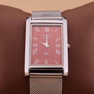 Op de afbeelding: Een rechthoekig Seiko Quartz horloge met een zilverkleurige kast en mesh band. De wijzerplaat is zalmkleurig met Romeinse cijfers en het Seiko Quartz logo. Het horloge wordt gepresenteerd op een bruin oppervlak.