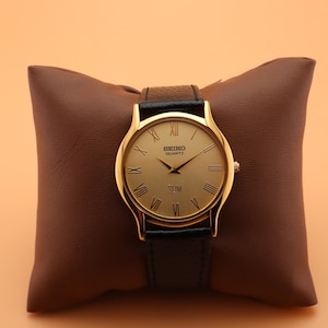 May include: A gold-toned Seiko Quartz Slim watch with a black leather band. The watch face features Roman numerals and the words "SEIKO QUARTZ SLIM". The watch is displayed on a brown pillow.