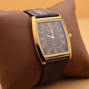 May include: A gold-toned Seiko Quartz watch with a rectangular black face and Roman numeral hour markers. The watch has a brown leather band and is displayed on a brown pillow.