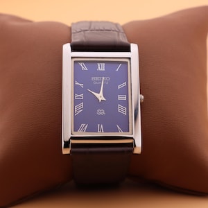 May include: A rectangular Seiko Quartz watch with a silver case and a dark blue face. The watch features Roman numeral hour markers and silver hands. The watch has a dark brown leather band and is displayed on a brown pillow.