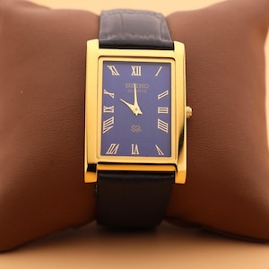 K&ouml;nnte beinhalten: Eine rechteckige Seiko Quartz-Uhr in Goldoptik mit blauem Zifferblatt und schwarzem Lederarmband. Die Uhr hat goldene r&ouml;mische Ziffern und Zeiger. Die Uhr wird auf einem braunen Lederkissen pr&auml;sentiert.