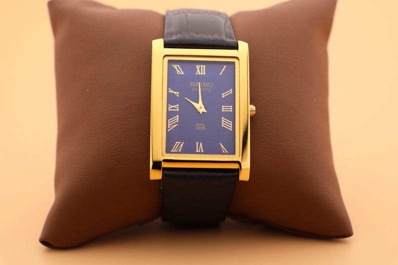 K&ouml;nnte beinhalten: Eine rechteckige Seiko Quartz-Uhr in Goldoptik mit blauem Zifferblatt und schwarzem Lederarmband. Die Uhr hat goldene r&ouml;mische Ziffern und Zeiger. Die Uhr wird auf einem braunen Lederkissen pr&auml;sentiert.