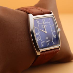 May include: A rectangular Seiko Quartz watch with a blue face and silver-tone case. The watch features Roman numeral hour markers and a brown leather strap. The watch is displayed on a brown leather pillow.