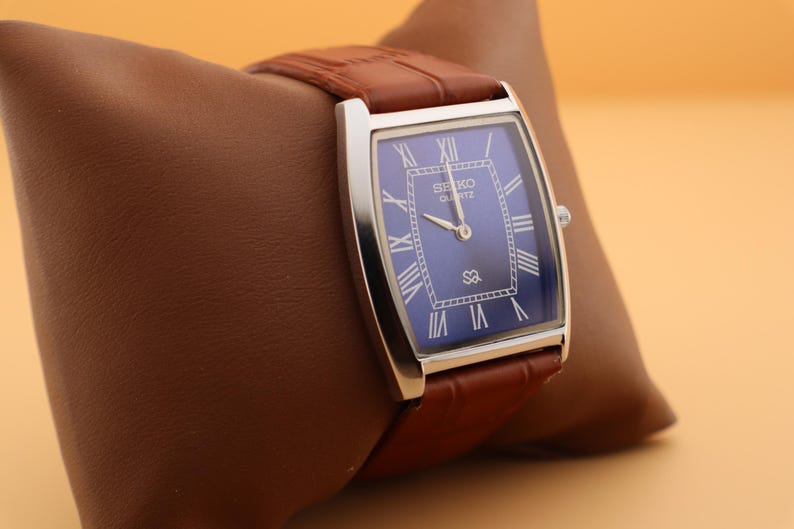 May include: A rectangular Seiko Quartz watch with a blue face and silver-tone case. The watch features Roman numeral hour markers and a brown leather strap. The watch is displayed on a brown leather pillow.