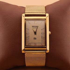 May include: A gold-toned Seiko Quartz watch with a rectangular face and mesh strap. The watch face features Roman numerals and the Seiko logo. The watch is displayed on a brown surface.