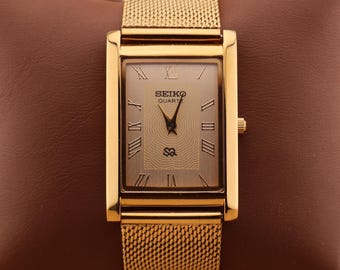 Può includere: Un orologio Seiko Quartz dorato con quadrante rettangolare e cinturino a maglia. Il quadrante dell'orologio presenta numeri romani e il logo Seiko. L'orologio è esposto su una superficie marrone.