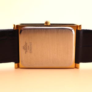 K&ouml;nnte beinhalten: Eine rechteckige Seiko-Uhr in Goldoptik mit einem schwarzen Lederarmband. Das Zifferblatt der Uhr ist geb&uuml;rstet goldfarben, und das Armband ist aus schwarzem Leder mit einer hellbraunen Unterseite. Die Uhr hat eine goldene Krone.