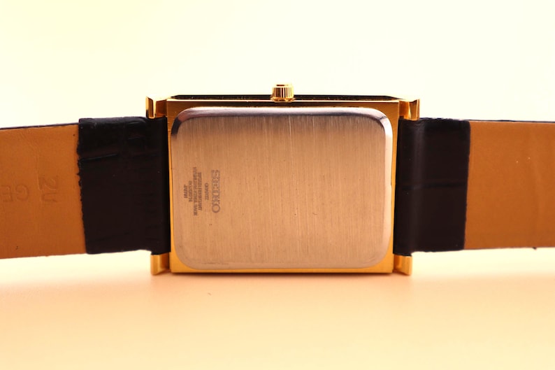 K&ouml;nnte beinhalten: Eine rechteckige Seiko-Uhr in Goldoptik mit einem schwarzen Lederarmband. Das Zifferblatt der Uhr ist geb&uuml;rstet goldfarben, und das Armband ist aus schwarzem Leder mit einer hellbraunen Unterseite. Die Uhr hat eine goldene Krone.