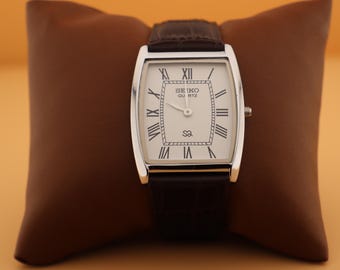 May include: A rectangular Seiko Quartz watch with a silver-colored case and a white face featuring black Roman numerals. The watch has a brown leather band and is displayed on a brown pillow.