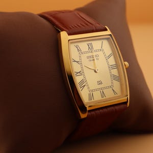 May include: A gold-toned Seiko Quartz watch with a rectangular face and Roman numeral hour markers. The watch has a brown leather band and is displayed on a brown pillow. The watch face displays the text "SEIKO QUARTZ".