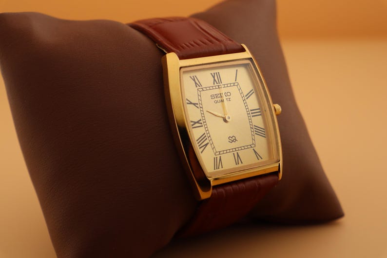 May include: A gold-toned Seiko Quartz watch with a rectangular face and Roman numeral hour markers. The watch has a brown leather band and is displayed on a brown pillow. The watch face displays the text "SEIKO QUARTZ".