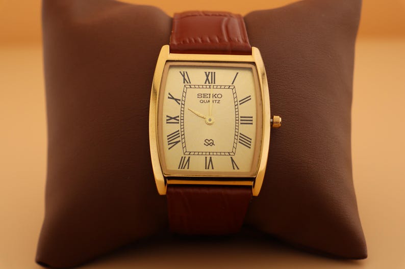 May include: A gold-toned Seiko Quartz watch with a rectangular face and brown leather strap. The watch face is cream-colored with black Roman numerals and the brand name. The watch is displayed on a brown leather pillow.