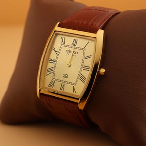 May include: A gold-toned Seiko Quartz watch with a rectangular face and Roman numeral hour markers. The watch has a brown leather band and is displayed on a brown pillow. The watch face displays the Seiko Quartz logo.