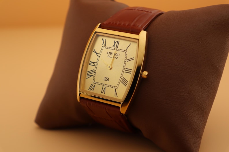 May include: A gold-toned Seiko Quartz watch with a rectangular face and Roman numeral hour markers. The watch has a brown leather band and is displayed on a brown pillow. The watch face displays the Seiko Quartz logo.