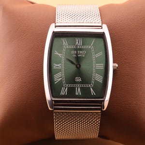 Könnte beinhalten: Eine silberne Seiko Quartz-Uhr mit einem rechteckigen grünen Zifferblatt und römischen Ziffern. Die Uhr hat ein Mesh-Armband und eine kleine Krone. Die Uhr wird auf einem braunen Kissen präsentiert.