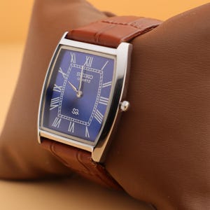 May include: A rectangular Seiko Quartz watch with a blue dial and silver-tone case. The watch features Roman numeral hour markers and a brown leather strap. The watch is displayed on a brown leather pillow.