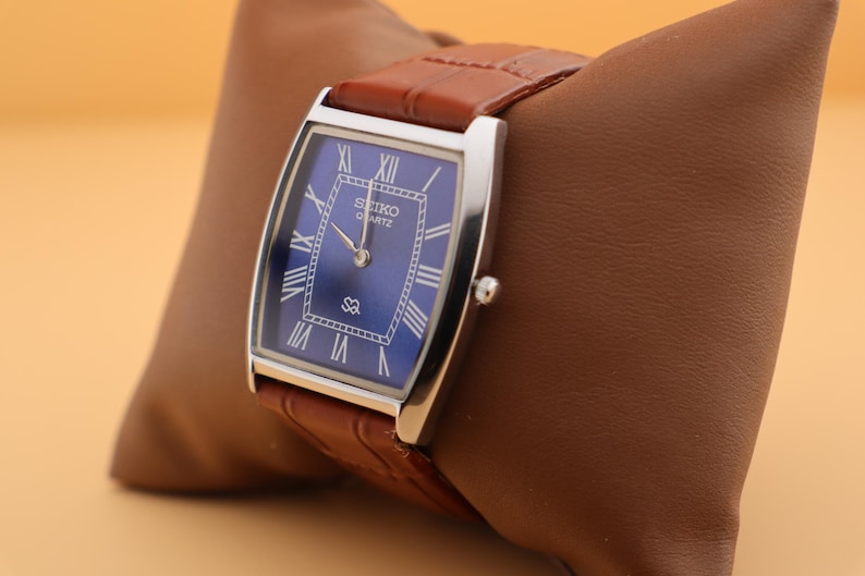 May include: A rectangular Seiko Quartz watch with a blue dial and silver-tone case. The watch features Roman numeral hour markers and a brown leather strap. The watch is displayed on a brown leather pillow.