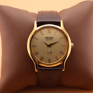 May include: A gold-toned Seiko Quartz Slim watch with a brown leather band, displayed on a brown leather pillow. The watch face has Roman numerals and black hands. The watch is a classic accessory.