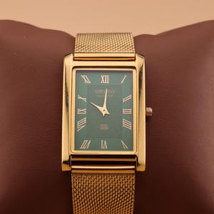 May include: A gold-toned rectangular Seiko watch with a green face and gold mesh band. The watch face features Roman numerals and a gold-colored hour and minute hand. The watch is displayed on a brown surface.