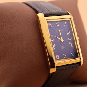 K&ouml;nnte beinhalten: Eine rechteckige, goldfarbene Seiko Quartz-Uhr mit dunkelblauem Zifferblatt und goldenen r&ouml;mischen Ziffern. Die Uhr hat ein schwarzes Lederarmband. Die Uhr wird auf einem braunen Kissen pr&auml;sentiert.
