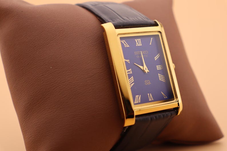 K&ouml;nnte beinhalten: Eine rechteckige, goldfarbene Seiko Quartz-Uhr mit dunkelblauem Zifferblatt und goldenen r&ouml;mischen Ziffern. Die Uhr hat ein schwarzes Lederarmband. Die Uhr wird auf einem braunen Kissen pr&auml;sentiert.