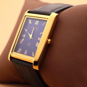 K&ouml;nnte beinhalten: Eine goldfarbene rechteckige Seiko Quartz-Uhr mit dunkelblauem Zifferblatt und schwarzem Lederarmband. Die Uhr hat goldene r&ouml;mische Ziffern und Zeiger. Die Uhr wird auf einem braunen Lederkissen pr&auml;sentiert.