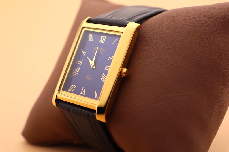 K&ouml;nnte beinhalten: Eine goldfarbene rechteckige Seiko Quartz-Uhr mit dunkelblauem Zifferblatt und schwarzem Lederarmband. Die Uhr hat goldene r&ouml;mische Ziffern und Zeiger. Die Uhr wird auf einem braunen Lederkissen pr&auml;sentiert.