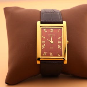 May include: A gold rectangular wristwatch with a burgundy face and black leather strap. The watch has Roman numeral hour markers and is displayed on a brown leather cushion.