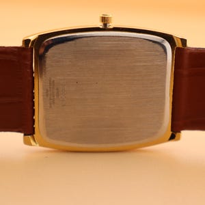 May include: Close-up of a gold-toned rectangular watch with a brown leather band. The watch face is silver, and the band is a rich brown color. The watch is set against a light orange background.