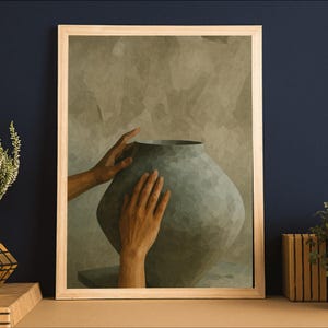 May include: A framed art print featuring a large, gray, textured vase with two hands gently touching it. The artwork is set against a muted, textured background. Decorative plants in gold pots and a Rubik's cube are on a wooden surface.