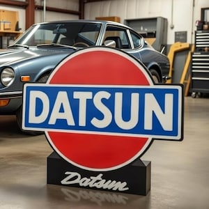 Op de afbeelding: Een klassieke blauwe Datsun sportwagen staat in een garage, met een groot Datsun-bord ervoor. Het bord heeft een rode cirkel en een blauwe rechthoek met het woord "Datsun" in het wit. De auto heeft een strak design met chromen accenten.