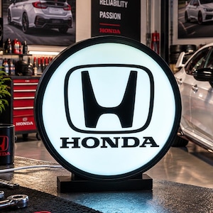 Puede incluir: Un letrero circular iluminado con el logotipo de Honda en negro sobre fondo blanco, colocado sobre un banco de trabajo oscuro. Al fondo, un Honda Civic Type R blanco está aparcado en un garaje con armarios de herramientas rojos y logos.