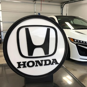 Op de afbeelding: Een rond Honda-logo bord met zwarte rand en het woord "HONDA" in zwarte letters. Een witte sportwagen is op de achtergrond te zien.