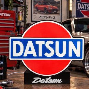 May include: A circular Datsun sign featuring a red background with a blue rectangular center containing the white text DATSUN. The sign sits on a black base with the Datsun logo in white script. The sign is illuminated and displays the brand name.