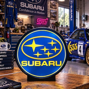 Könnte beinhalten: Ein rundes, beleuchtetes Subaru-Schild mit dem blau-gelben Sternenlogo und Schriftzug der Marke. Das Schild steht auf einer Werkbank aus Holz in einer Werkstatt, im Hintergrund sind ein blaues Subaru-Rallyeauto, Werkzeuge und Fanartikel zu sehen.