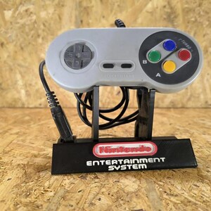 Può includere: Un controller per videogiochi grigio con un'area pulsanti circolare nera, un pad direzionale grigio e quattro pulsanti azione colorati etichettati A e B. Il controller è montato su un supporto nero con il logo Nintendo Entertainment System.