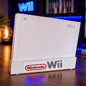 Może przedstawiać: Biała konsola do gier wideo Nintendo Wii ustawiona pionowo na dopasowanej białej plastikowej podstawce. Konsola posiada logo Nintendo oraz oznaczenie ATI z boku, natomiast na podstawce widnieje czerwone logo Nintendo i szary napis Wii.