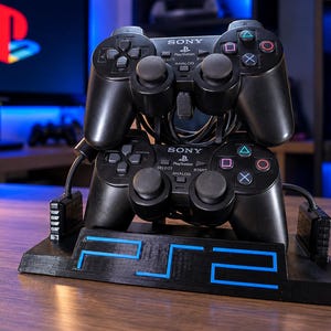 Op de afbeelding: Twee zwarte Sony PlayStation 2-controllers gestapeld op een zwarte displaystandaard. De standaard is aan de voorzijde voorzien van de blauwe tekst PS2. Elke controller heeft twee analoge sticks, een D-pad en vier actieknoppen.