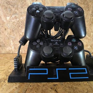 Peut inclure: Deux manettes de jeu Sony PlayStation 2 noires montées sur un support d'exposition noir. Le support présente le logo PS2 bleu sur la base avant. Chaque manette possède deux joysticks analogiques, une croix directionnelle et quatre boutons.